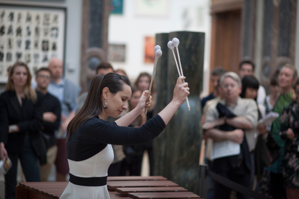 A female student, dressed in a black and white top, holding a pair of mallets in a xylophone, performing in front of a crowd.